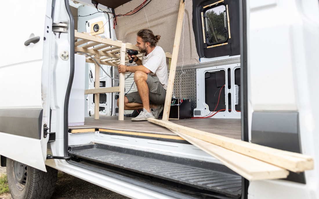 Image of a man building shelves inside a van with a drill, surrounded by wood and tools, converting it for van life.