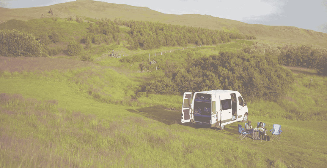 A white camper van parked in a scenic grassy field with hills in the background, showing two people sitting at a folding table with chairs, enjoying van life—perfect for exploring how much is a camper van.