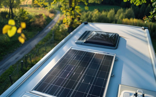 A solar panel on a camper van roof with a vent, overlooking a scenic landscape with trees, a winding road, and mountains under a clear sky.