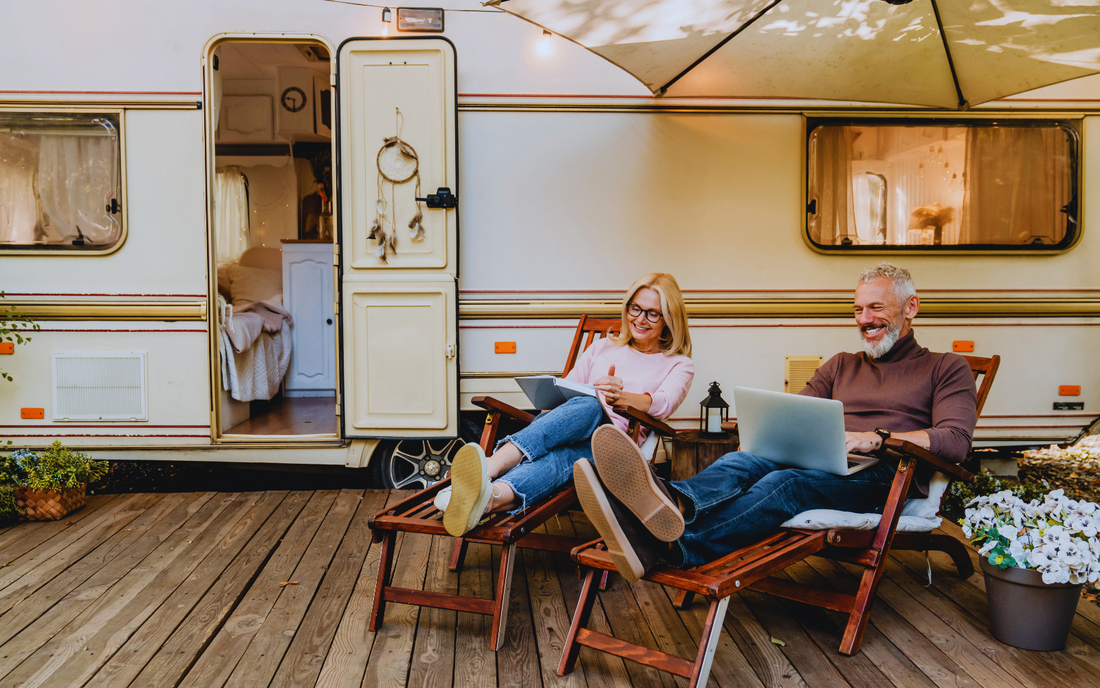 An older couple relaxing on chairs outside a caravan, using laptops, with a wooden deck, plants, and a scenic setup under a canopy.
