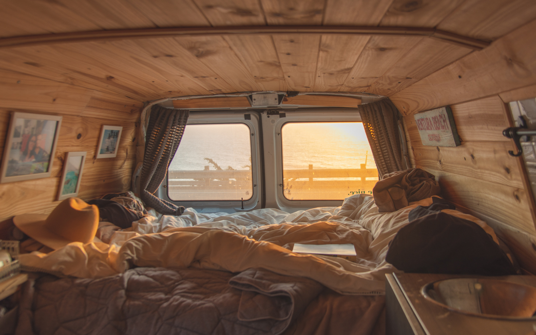 Interior view of a van with wooden walls, a bed with blankets, a cowboy hat, and photos, overlooking a sunset sea view from Ventura Beach, California.