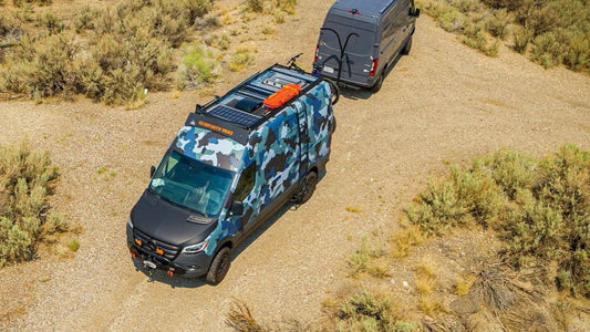 Aerial view of two Mercedes Sprinter vans parked on desert dirt road - one with blue camouflage wrap and solar panels, one grey - showcasing van life adventure travel
